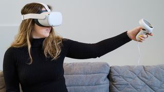 Wanita muda mencoba Meta Quest 2 (Oculus Quest 2) untuk pengalaman virtual yang duduk di sofa, mengenakan atasan hitam