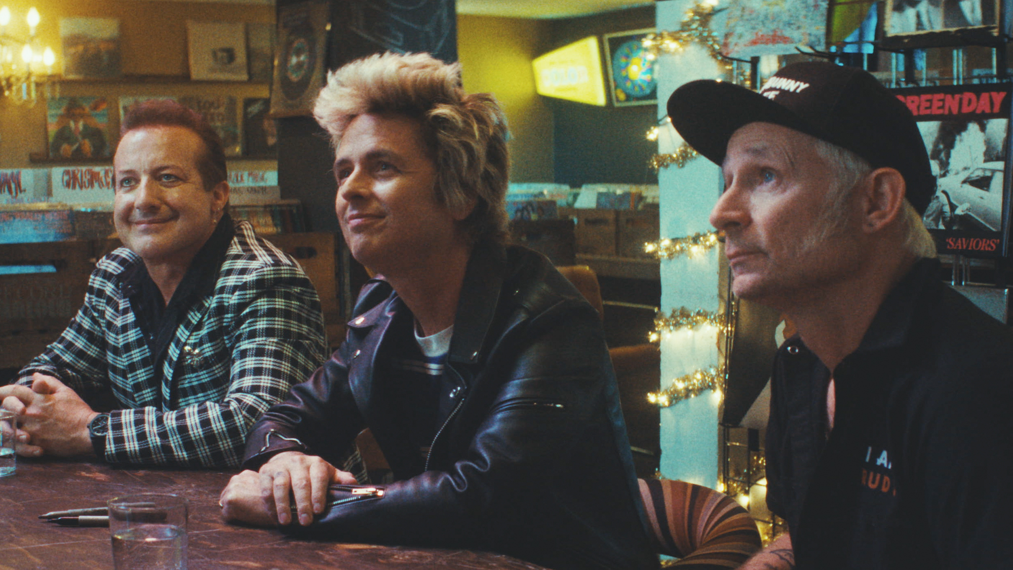 Green Day sitting at a table, looking up. From left to right, Tre Cool, Billie Joe Armstrong, Mike Dirnt