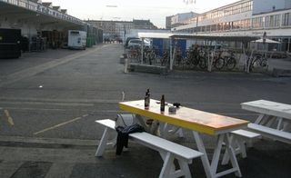 A factory building with a bike shed and long tables with benches next to it in front of a road with a bridge on the other side of it.