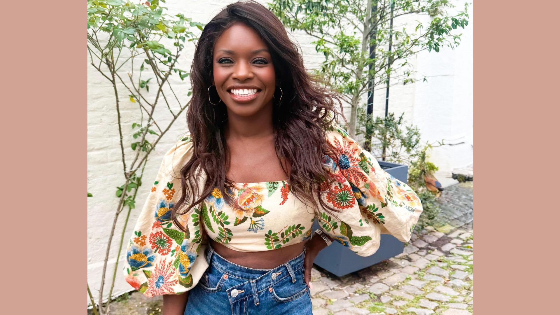 A smiling woman with long, wavy hair wearing a colourful floral top and high-waisted jeans, standing in a cobblestone courtyard, reflecting themes of beauty and personal transformation.