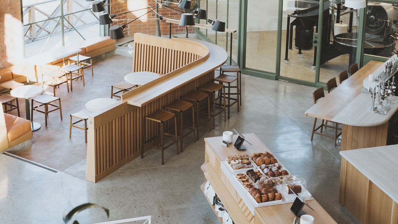 A sun-filled, minimalist coffee shop features light wooden furniture in clean, playful lines, green iron doors, and plenty of pastries.