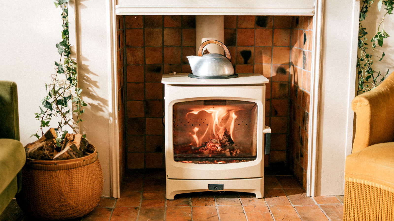 Cream log burner with kettle on top in tiled fireplace with plants and chairs either side