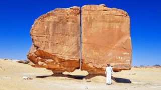 A person dressed in white stands in front of the Al Naslaa Rock Formation. The formation consists of two stone blocks on small pedestals with a fine, perfectly straight gap between them.