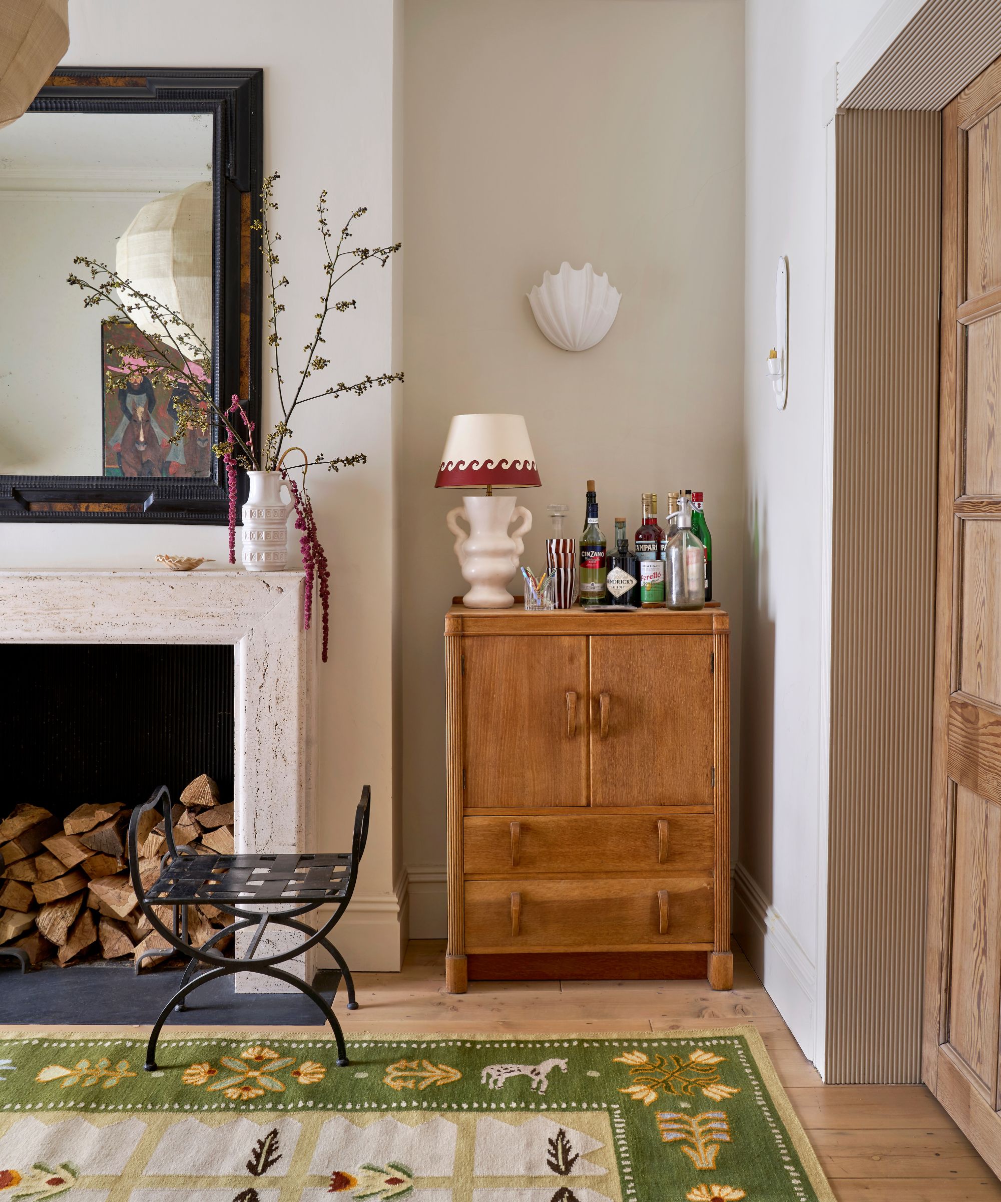 neutral &#039;middle room&#039; in a traditional victorian home with a large stone fireplace, a small wooden vintage cabinet styled with a lamp, a large overhead ceiling pendant, small shell wall sconces and a folk floral rug