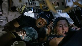A man and a woman hunch over in a small capsule, both looking at separate electronic screens