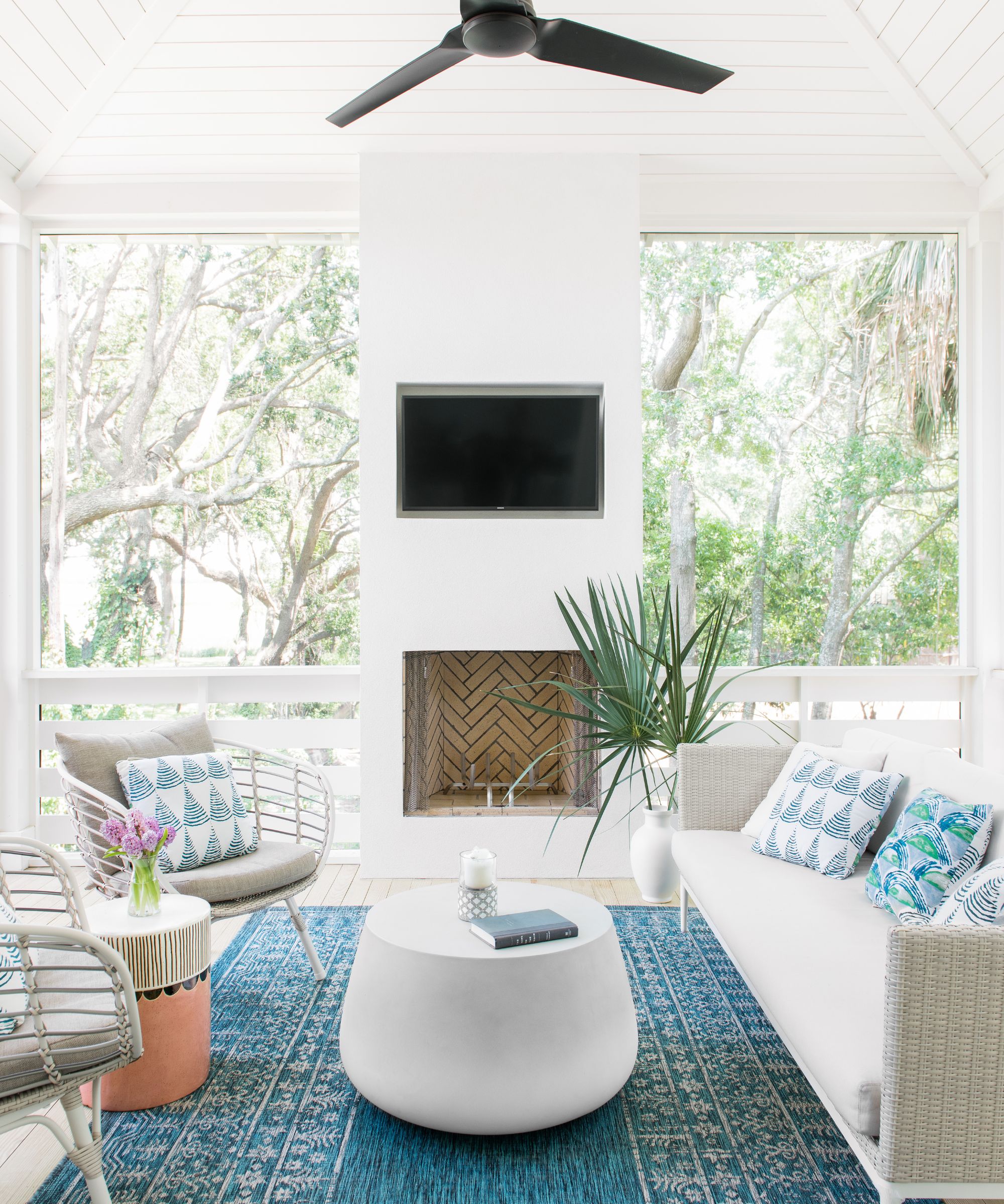 Sunroom with wood panelling painted white, fireplace, and blue rug with sofa and rattan chairs and round coffee table