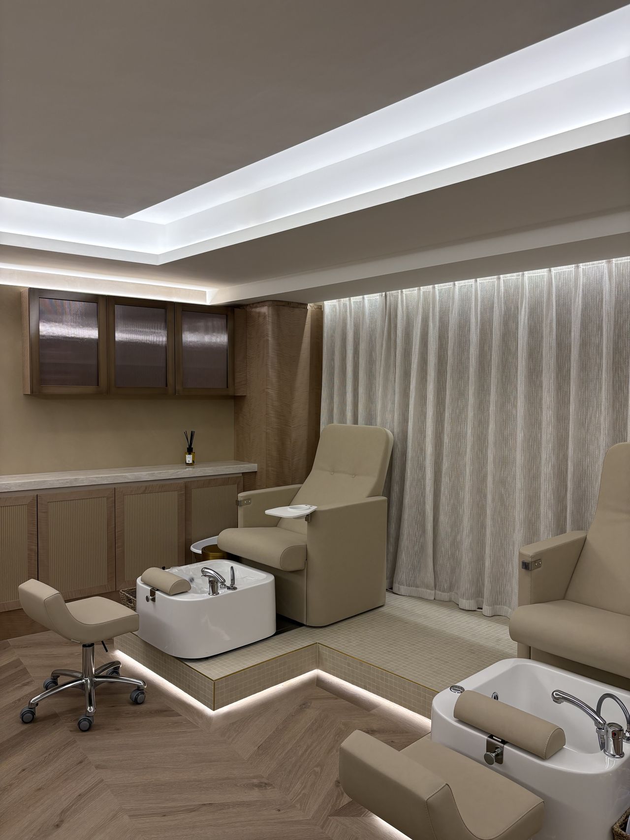 A beige-tinted beauty salon features matching leather armchairs for treatments, wheels-equipped stools, wooden furniture, and floors, all brought to life by dramatic ceiling lighting and flowy white curtains.