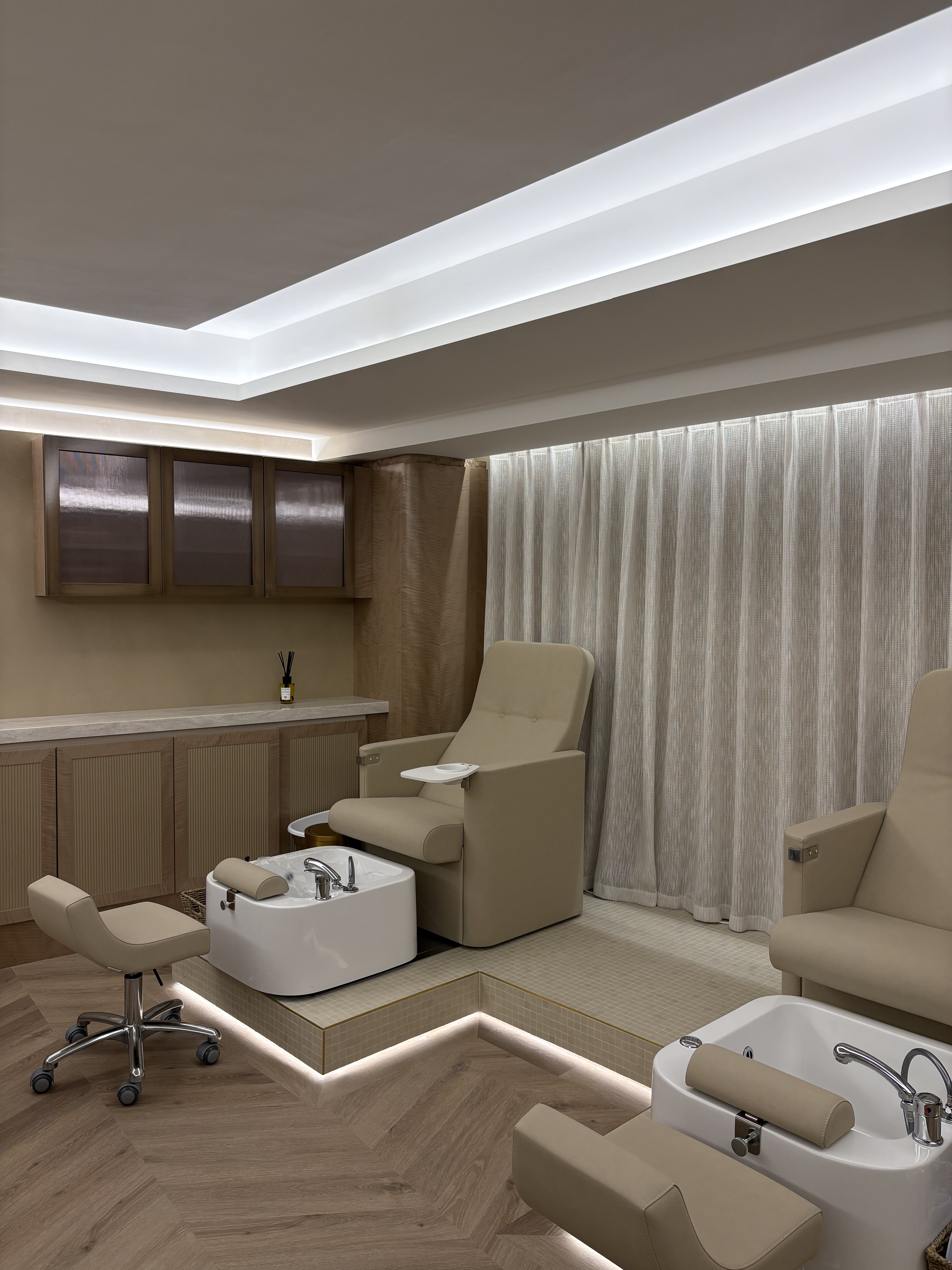A beige-tinted beauty salon features matching leather armchairs for treatments, wheels-equipped stools, wooden furniture, and floors, all brought to life by dramatic ceiling lighting and flowy white curtains.