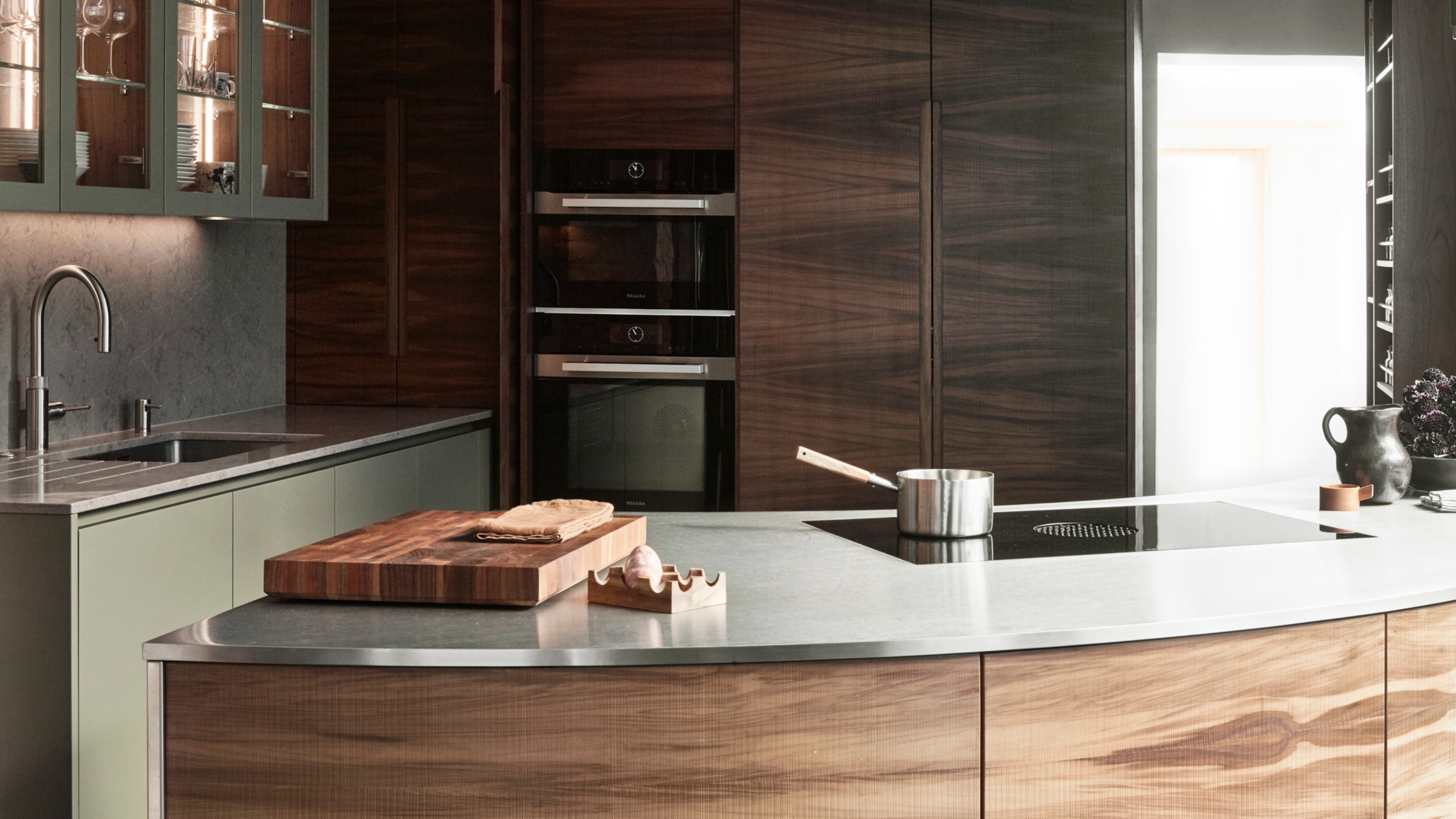 a modern kitchen with stainless steel counters, wooden cabinetry and sage green cupboards. The kitchen island is curved and separates the kitchen