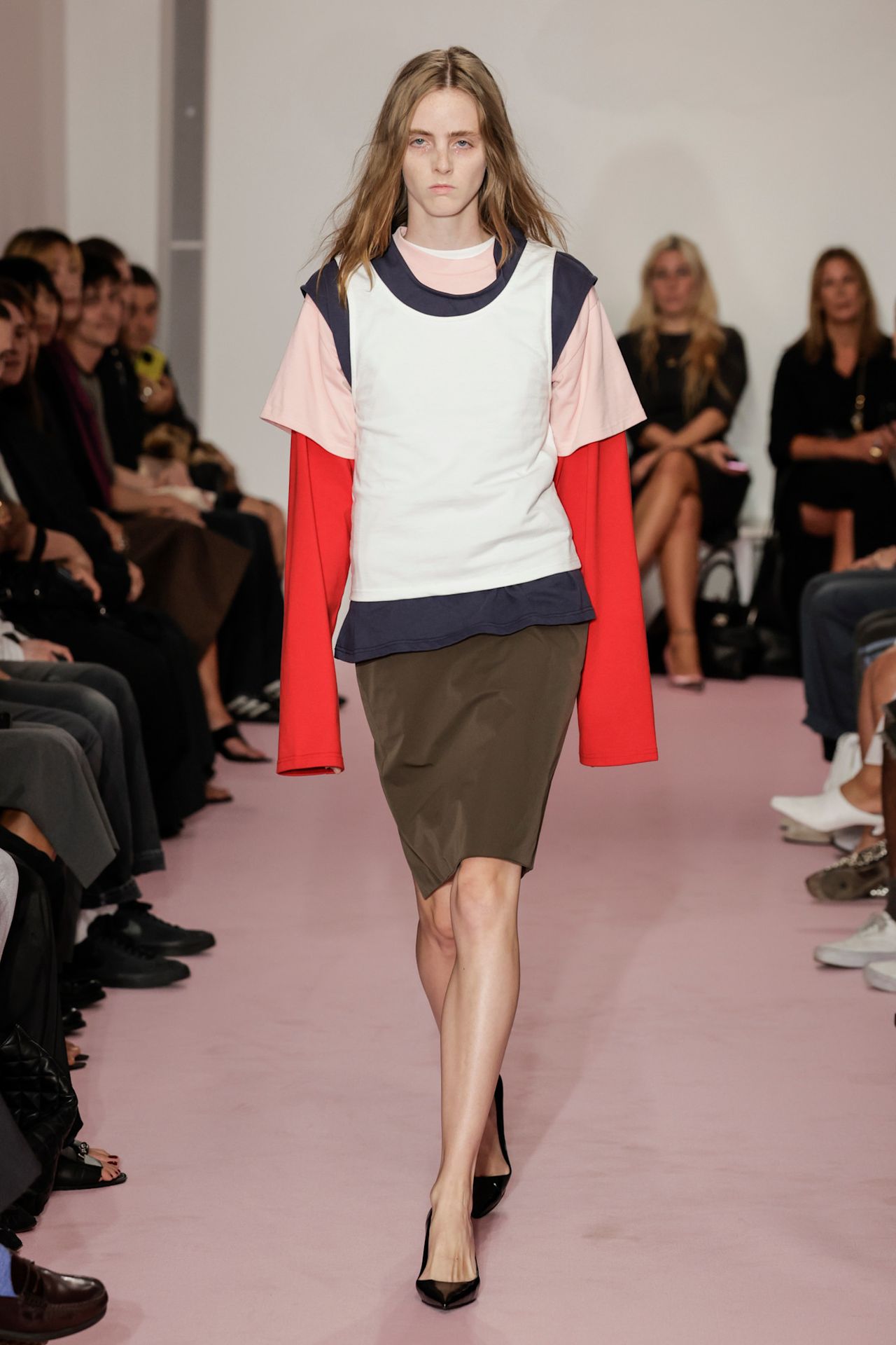 Layering staples for spring 2026 is shown in a photo from LII's runway show, with a model walking while wearing an oversized red button-down shirt layered under a light pink s-shirt, a blue t-shirt, and a white tank top. The layers are paired with a brown pencil skirt and black pointed pumps