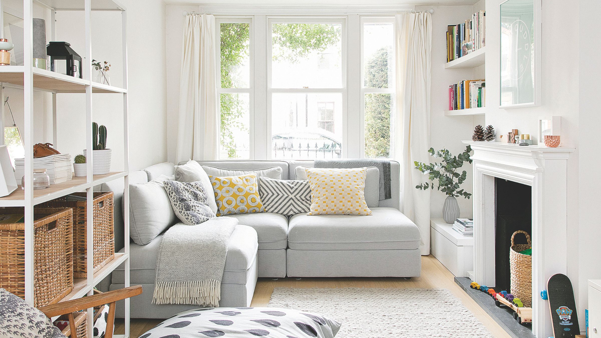 White painted living room with a grey sofa in front of a window, and storage opposite a white fireplace