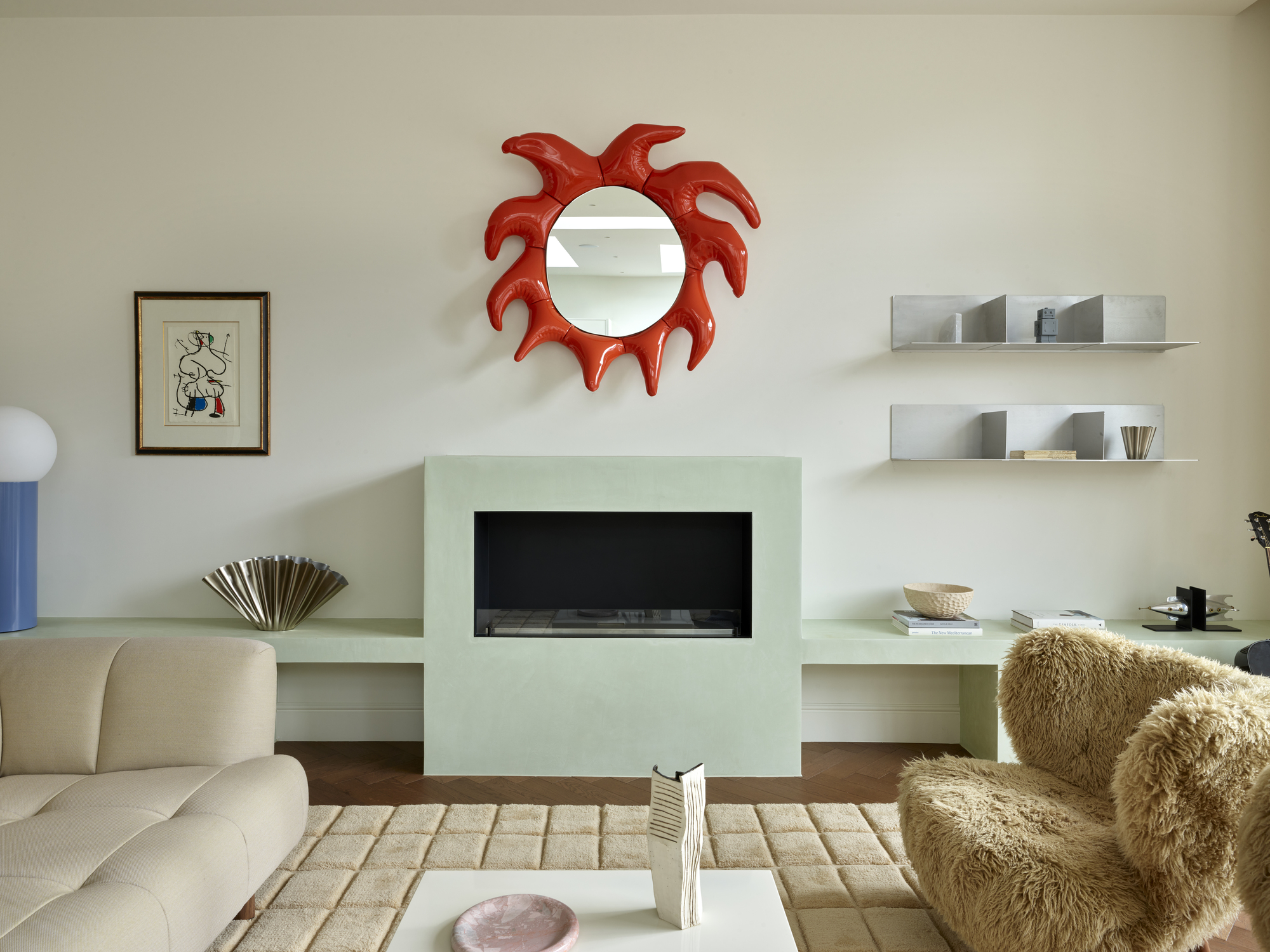 Image of an off-white living room wall with a modern, light mint green fireplace. There is a sculptural, red mirror over the fireplace, a fuzzy accent chair, and chrome shelving in the room.