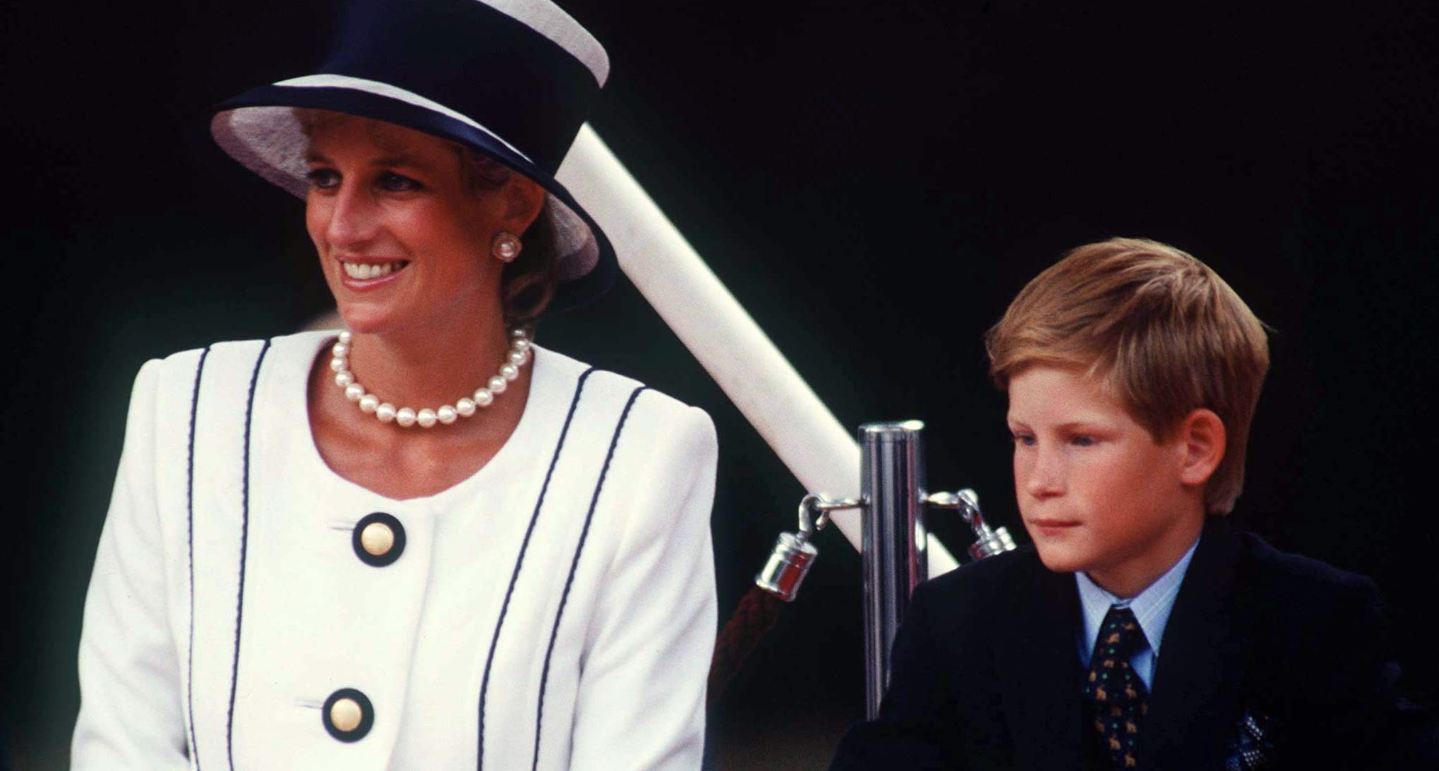 Prince Harry sitting next to Princess Diana, wearing a white blazer and blue hat