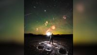 A person standing on a rock looks up at a green night sky with streaks of light showing a meteor shower overhead