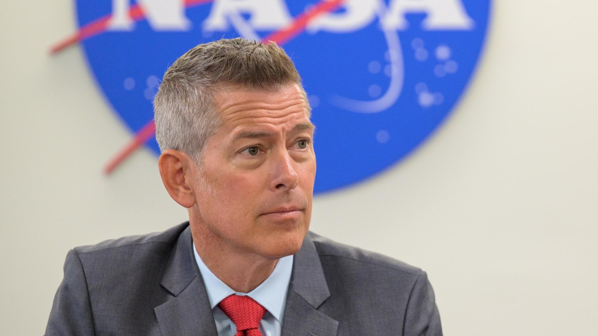 A man in a suit sit looks to the right in front of the bottom half of a nasa meatball logo.