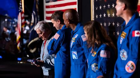 four people in blue jumpsuits from the right, look left and a grey-haired man in a light blue jacket holding a folded american flag.