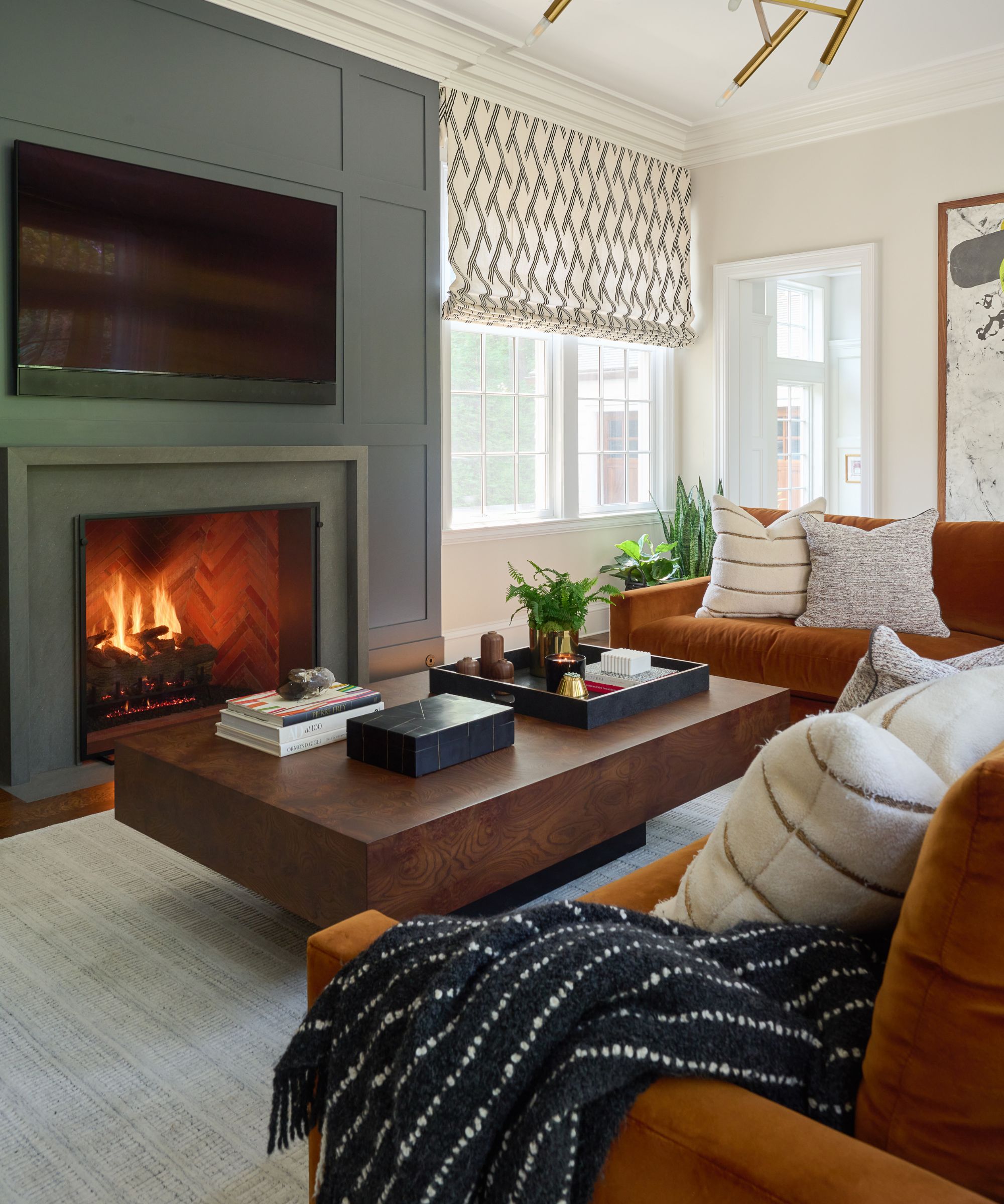 green and orange cozy modern living room with a fireplace panelled and painted an earthy green, lit fire, wood coffee table and a large rust orange velvet couch