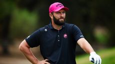 Jon Rahm wearing a pink cap and black polo while resting his hand on his driver