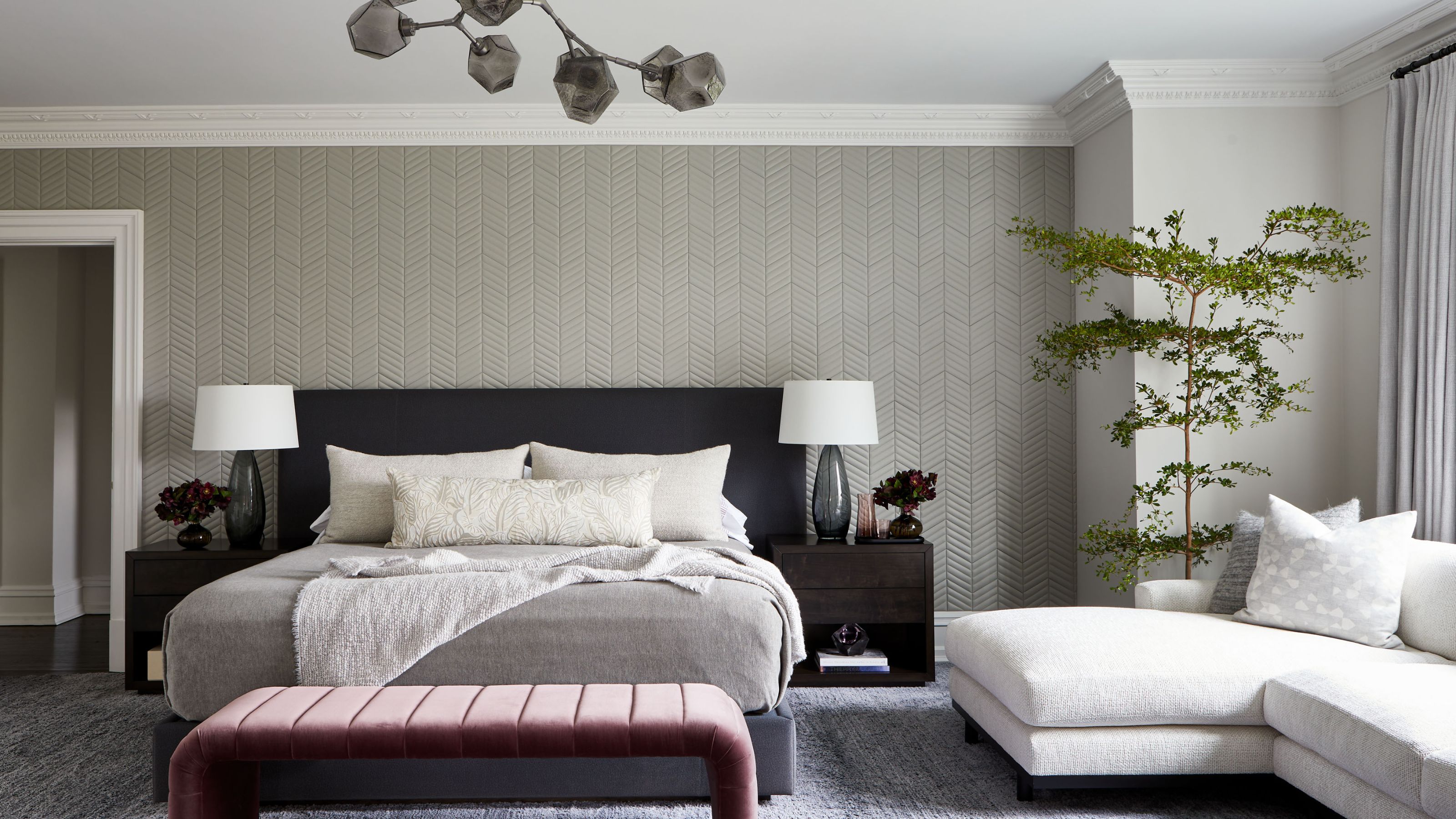 Large master bedroom with gray upholstered leather walls and a large bed with dark gray headboard. A pink velvet bench is at the end of the bes and a cream sectional sofa is under the window