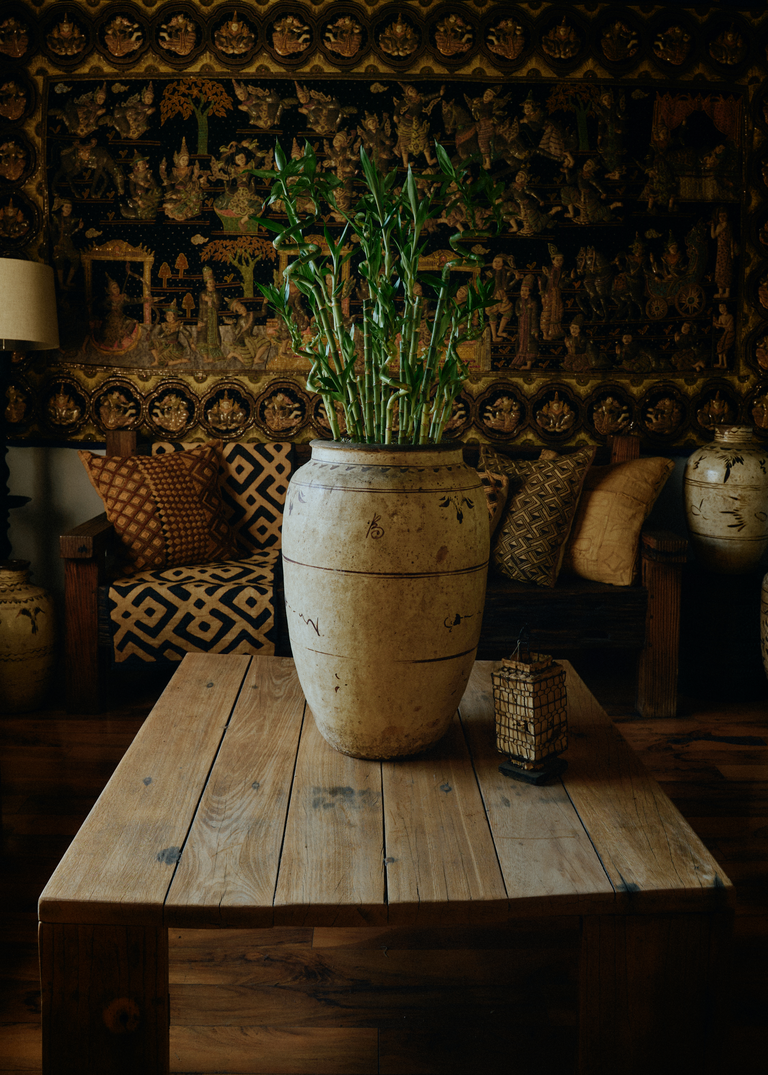 an image of lucky bamboo spirals styled in a large vessel on top of a rustic wooden coffee table in front of a sofa with lots of patterned cushions and a dark, detailed wall mural behind it with a lamp shade in one corner and an empty vessel in the other corner