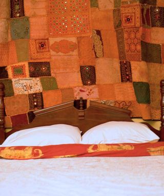 An orange and brown patchwork tapestry hanging above a dark brown wooden bed with white sheets with an orange trim
