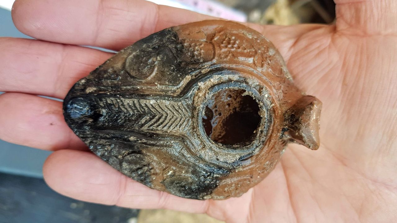 A small pottery oil lamp held in someone&rsquo;s palm. It is black and brown, with some simple markings on it, such as circles and arrows.