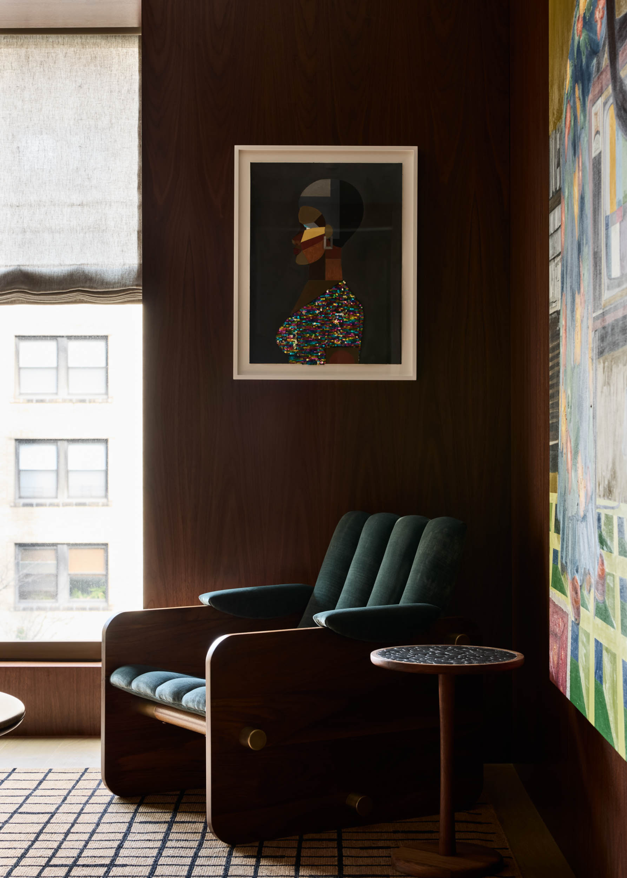 A brown living room with wood walls, a framed painting, a ribbed dark teal chair and a marble top side table by a large abstract colorful painting