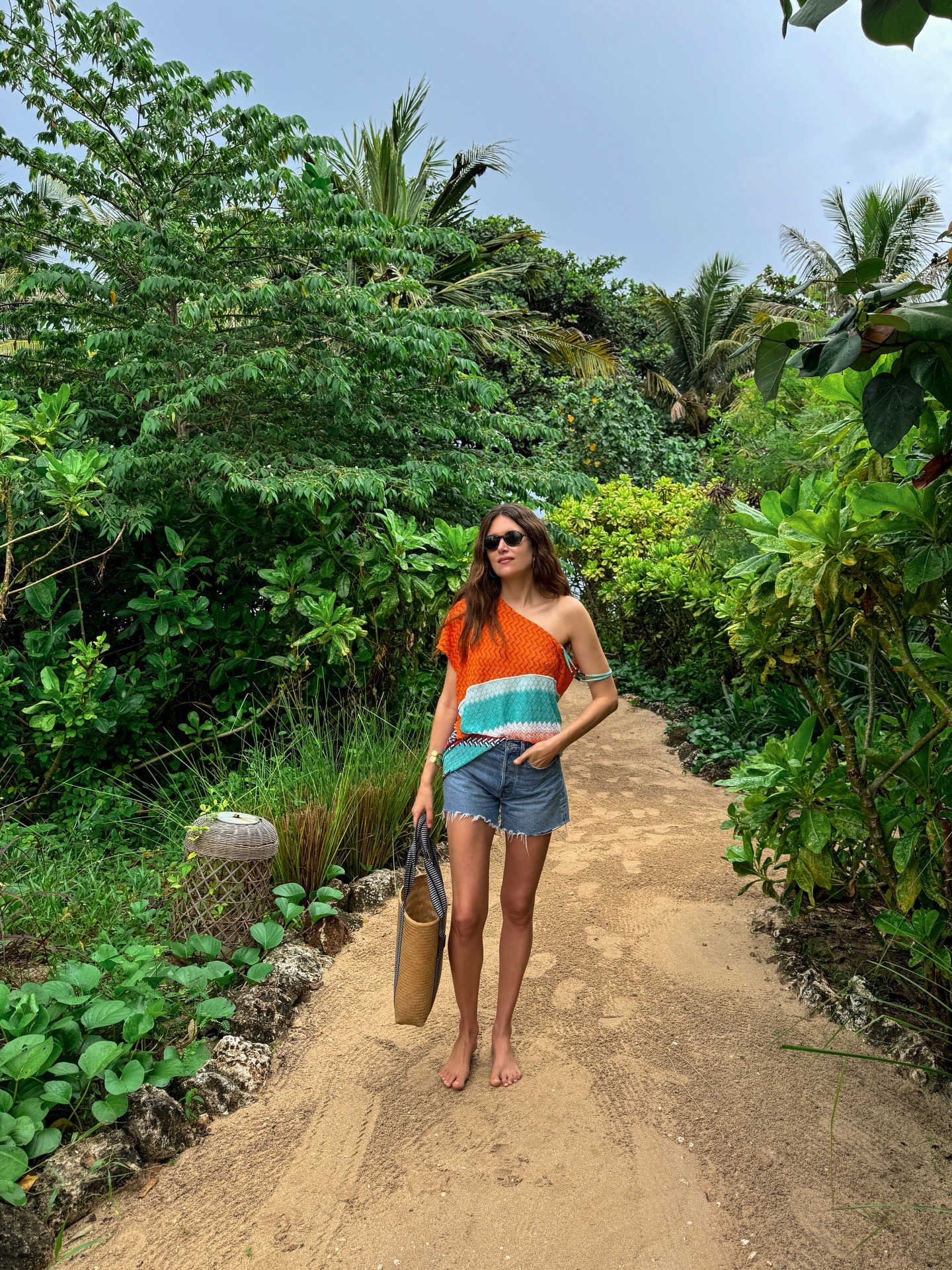 Fashion person Sara Loura wears a winter sun outfit. She wears denim short a one shoulder top and carries a raffia bag. She is walking barefoot in a recent image taken from her Instagram.