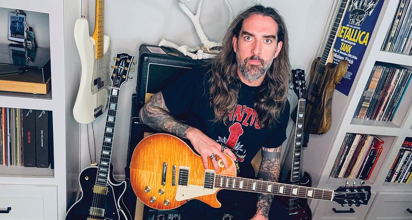 Nate Garrett of Spirit Adrift pictured at home with a Gibson Les Paul. He wears a Danzig T-shirt and is there are books, amps, and LPs in the background.