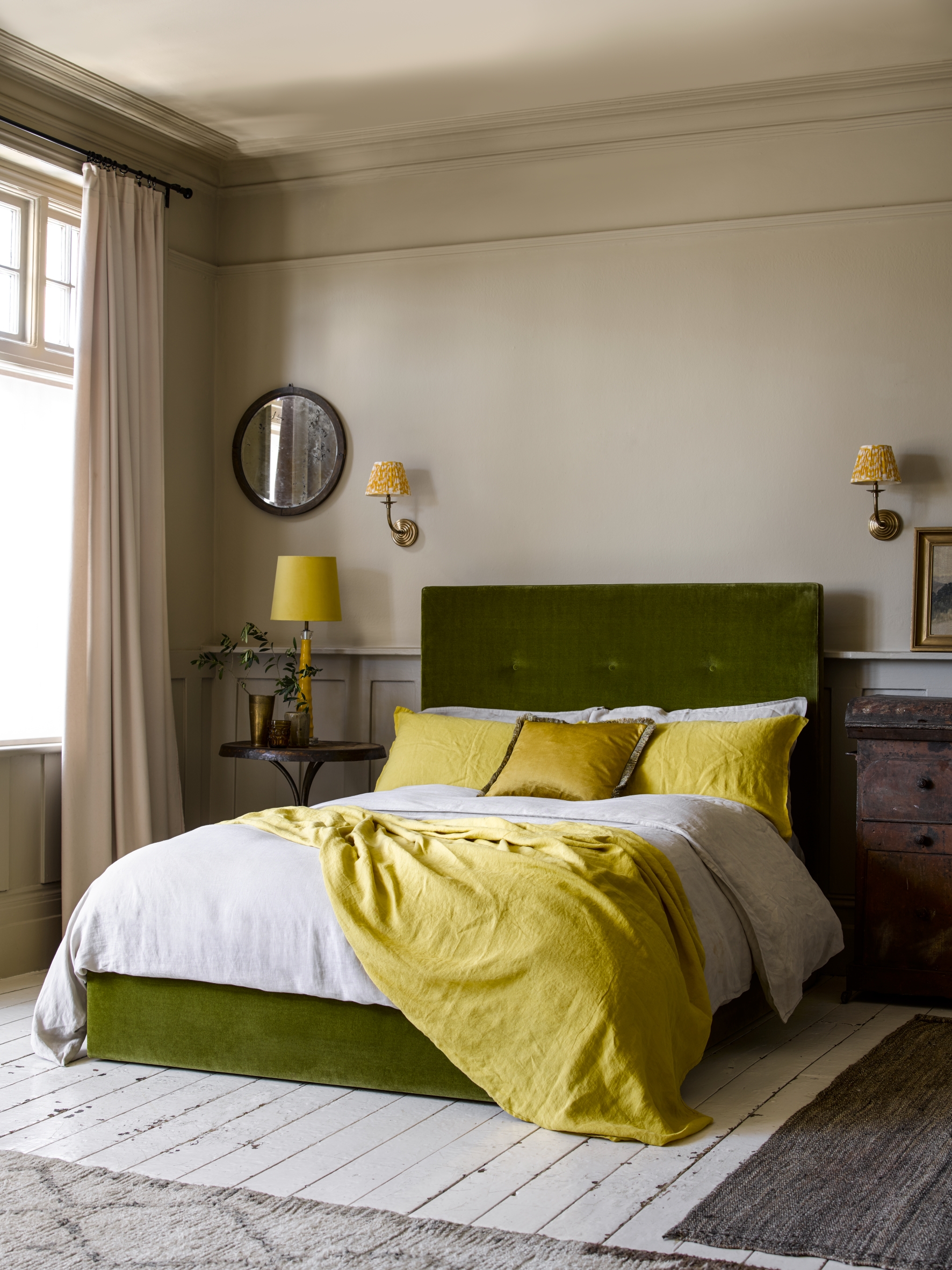 The best bedroom colors in a neutral beige scheme, with a green velvet upholstered bed, white floorboards and bright citrus yellow lighting, pillows and throw.