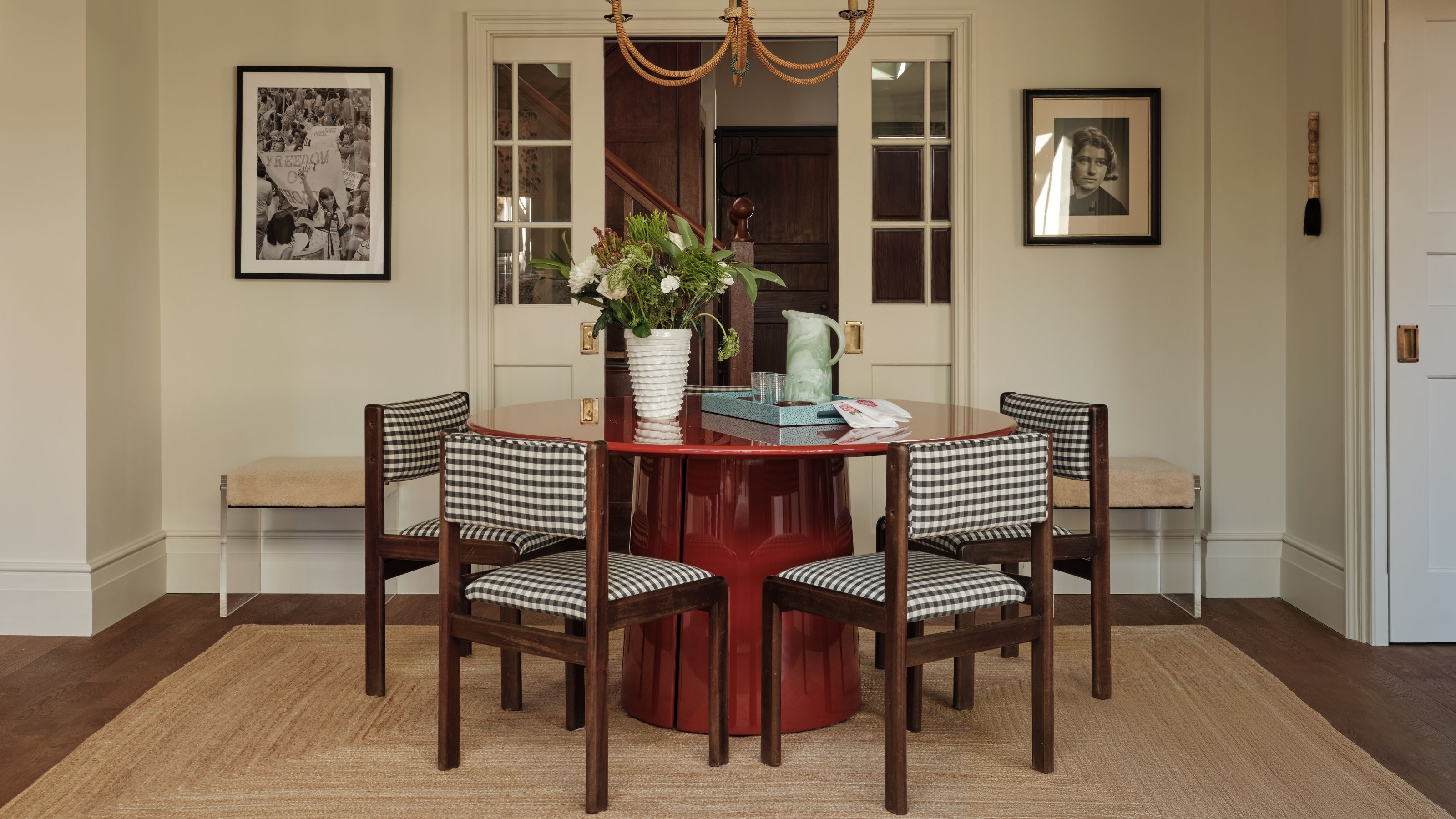 Image of a white dining room with a round, glossy red dining table with five black and white gingham dining chairs around it. There is a jute rug underneath the table.