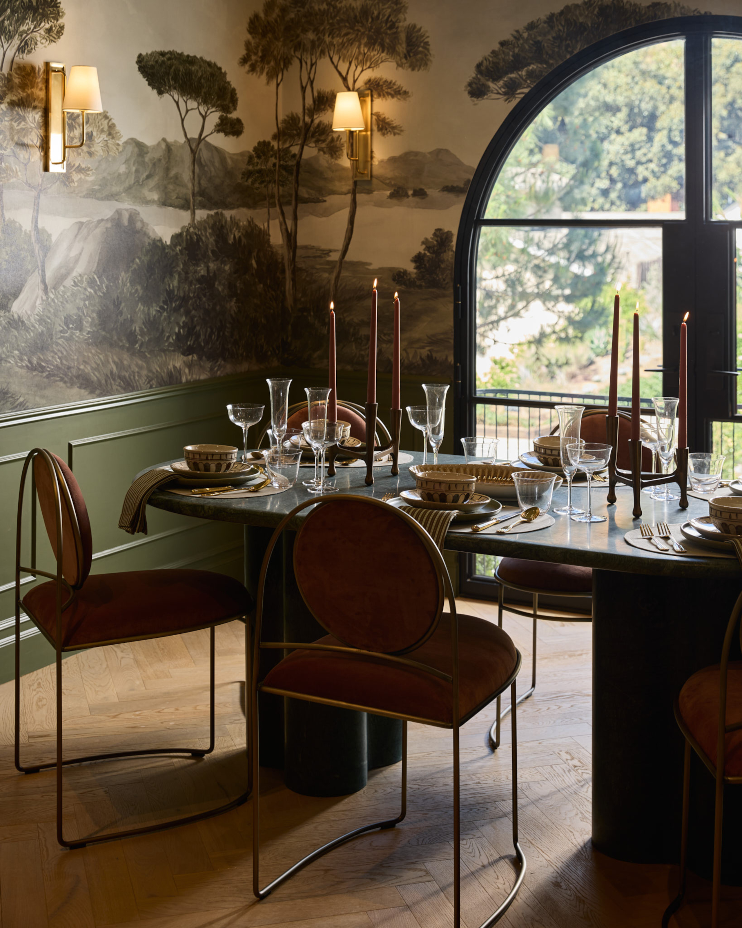 Dining area with green panelled walls and wallpaper depicting trees, dark green dining table with marble top, rust-colored velvet chairs, gold wall sconces and red candlesticks