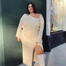 Plus size influencer Gia Sintra wearing off-white asymmetrical knit dress with white thigh-high boots, standing in front of a greeen wall. 