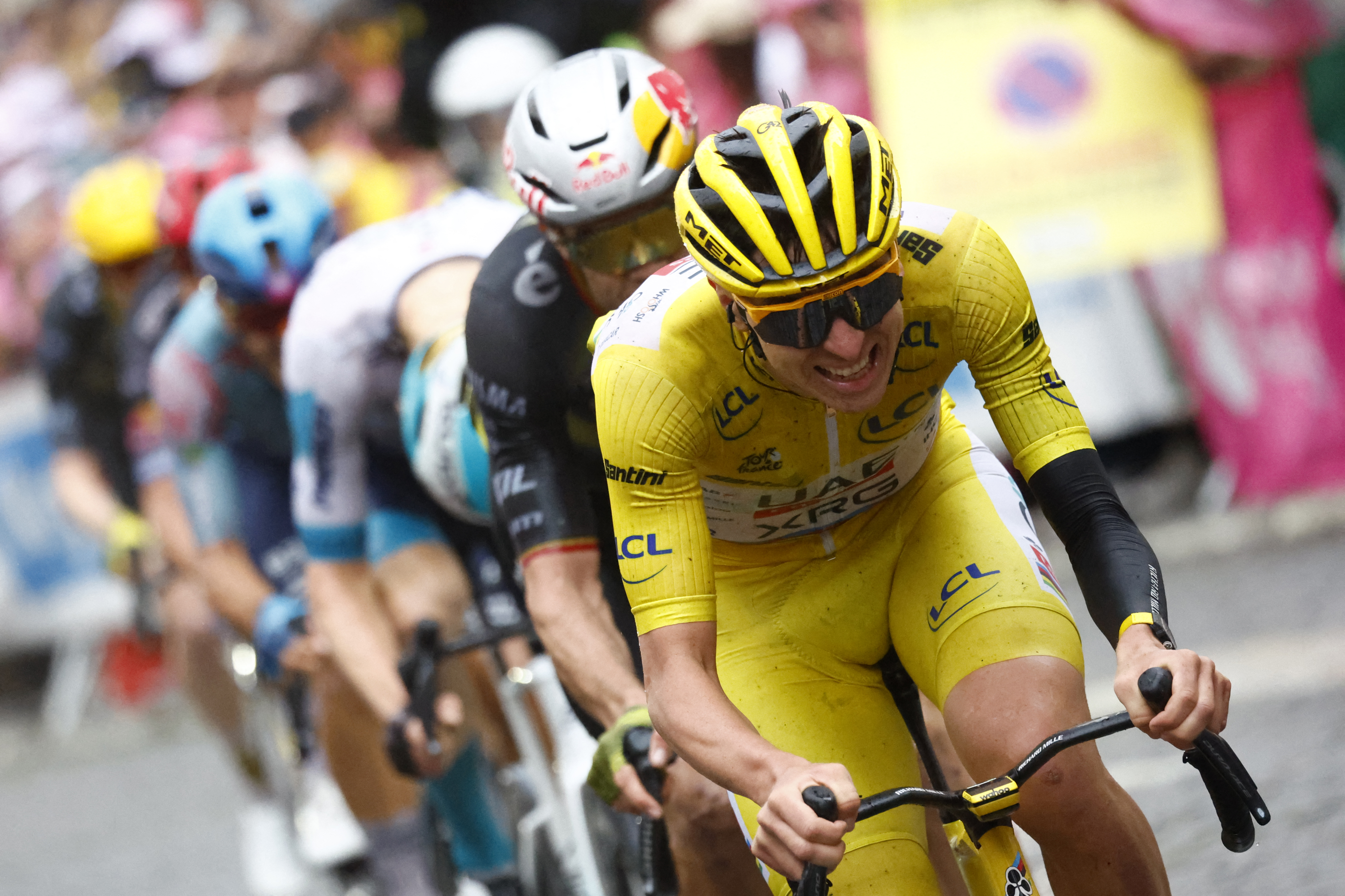 Tadej Pogačar riding in the Tour de France wearing the yellow leader's jersey, grimacing as he leads a line of riders including Wout van Aert