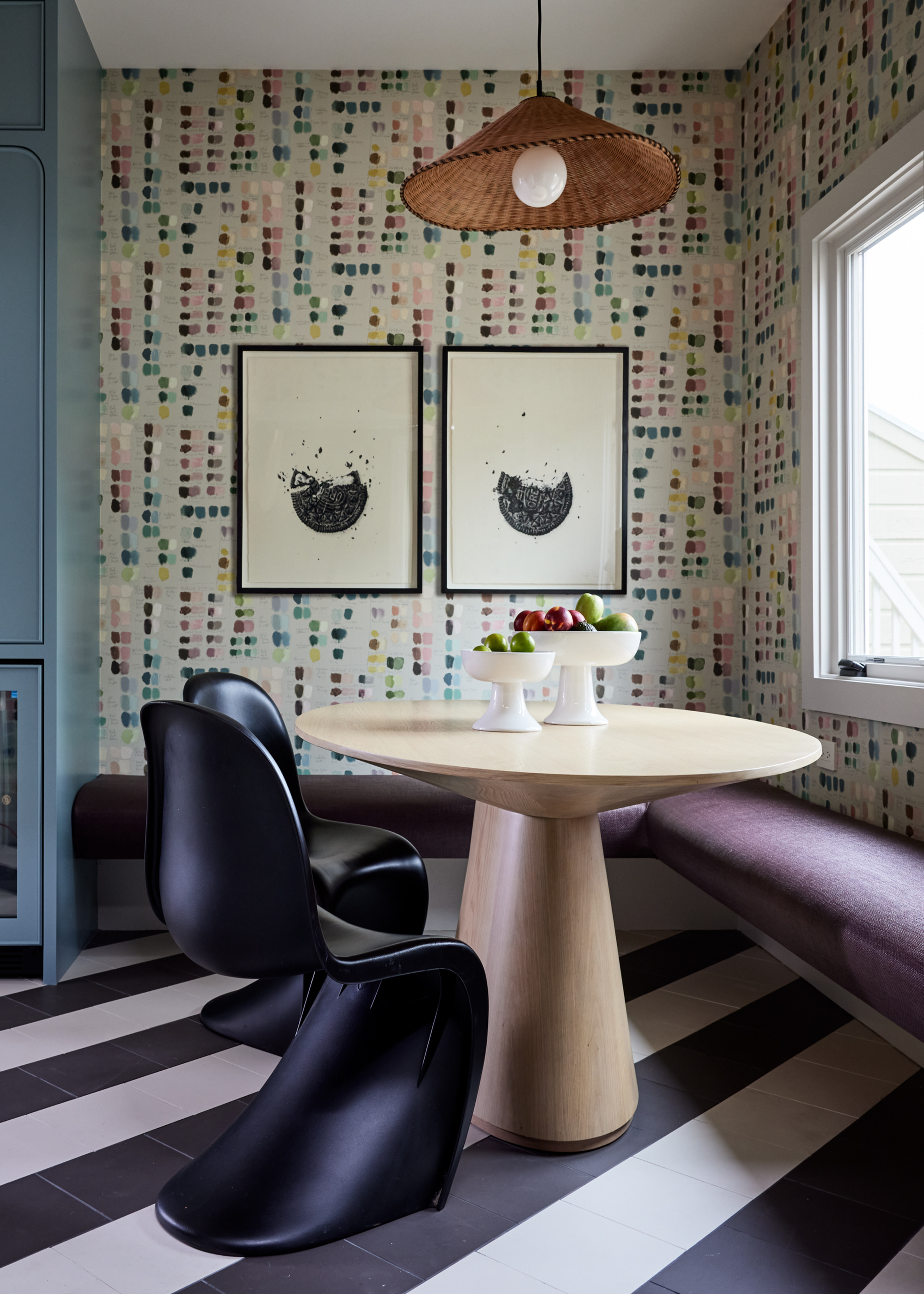 A small circular dining table with two sculptural chairs and a banquette bench with two bowls of fruit in the center