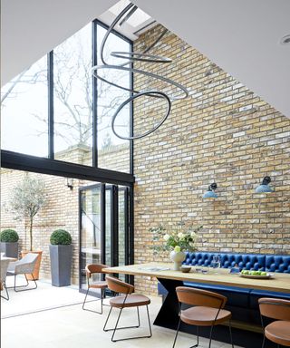 Large kitchen with double height ceilings, artistic hanging light, exposed brick wall, large wooden dining table with metal base, blue booth seating, brown leather and black dining chairs, large glass windows and doors opening out onto garden