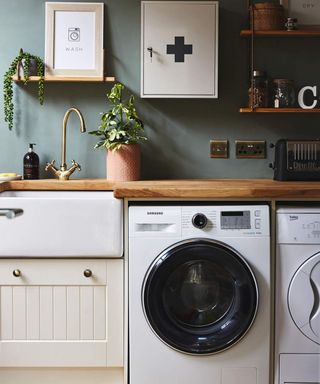 A kitchen with dark green wall paint decor, wooden shelves, brass-colored kitchen faucet, Samsung washing machine, Beko dryer and Dualit toaster