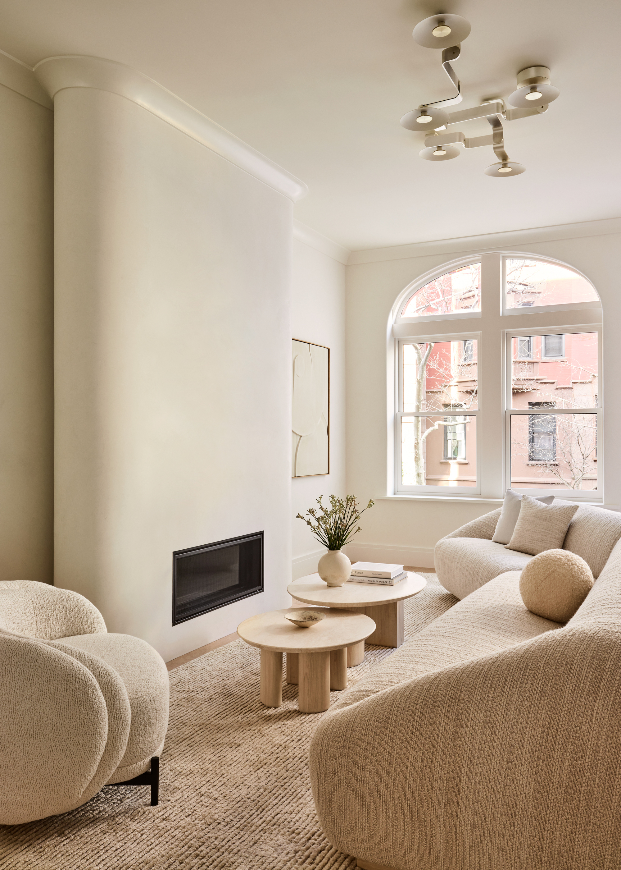 beige living room with curved beige sofa, arched window, pendant ceiling light, a curved architectural fireplace, two nesting travertine tables, and a cream armchair