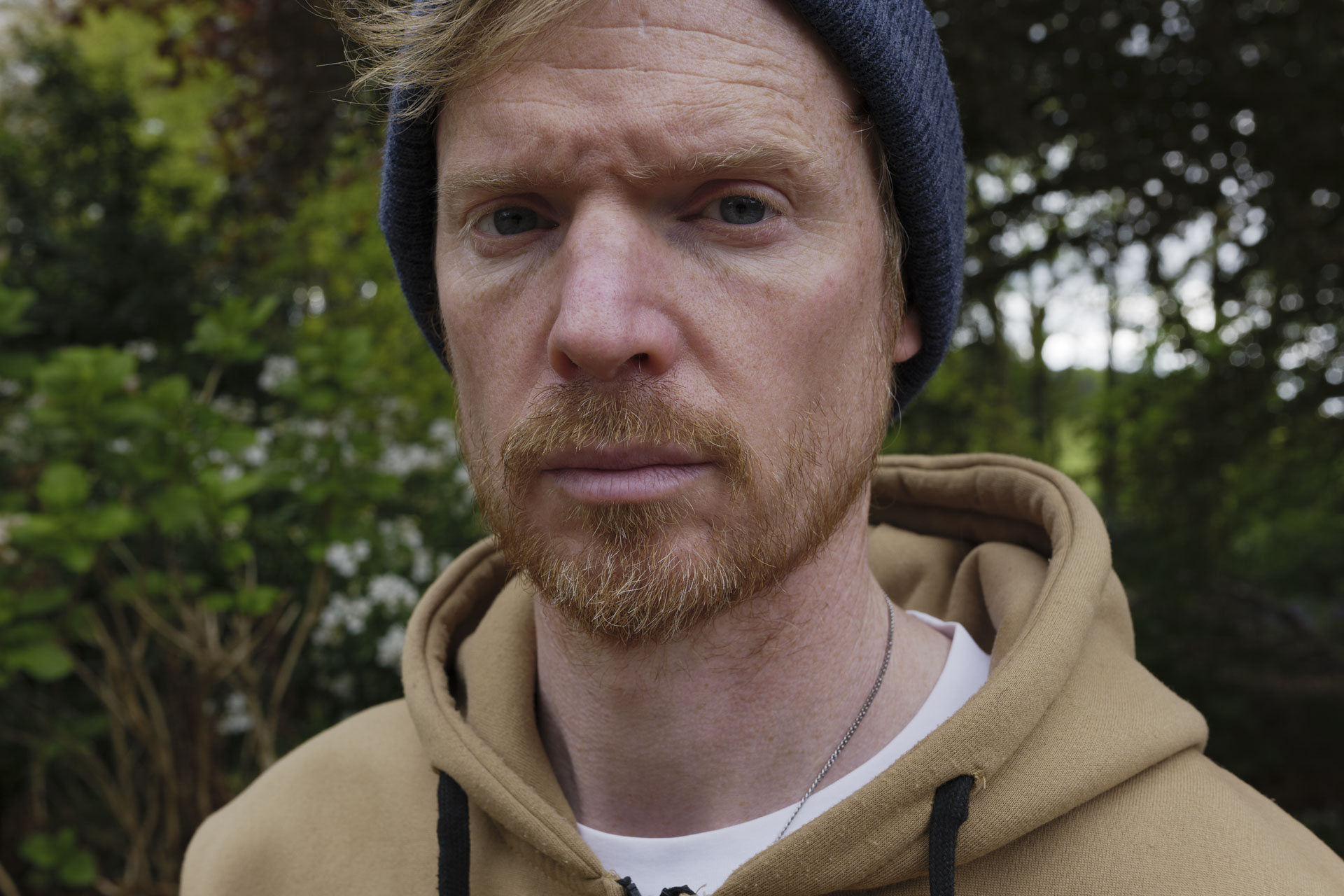 Portrait of a man in a beanie hat and hoodie, the background is lush green bushes and trees with dappled light