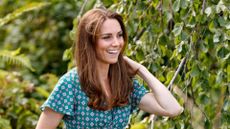 Kate Middleton smiling as she stands outdoors near some trees and plants