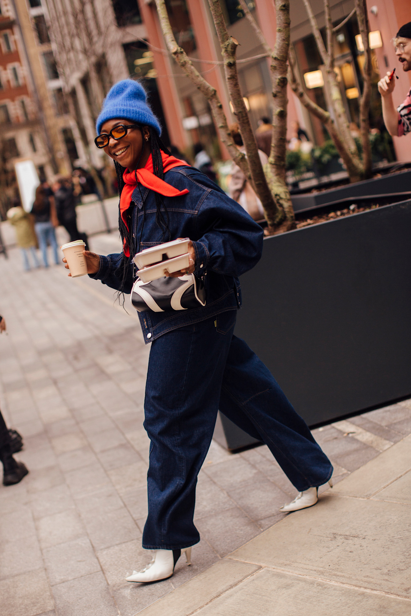a woman at london fashion week wearing a red triangle scarf, denim jacket, jeans, white boots, and a blue beanie hat