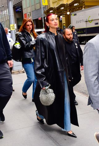 Dua Lipa in New York City April 2024 wearing the leather trench coat trend and carrying a silver bag