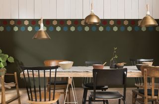 Trio of brass pendants suspended over trestle style table, with half and half walls featuring dark painted section with colored hexagons border, and white paneling on upper section.