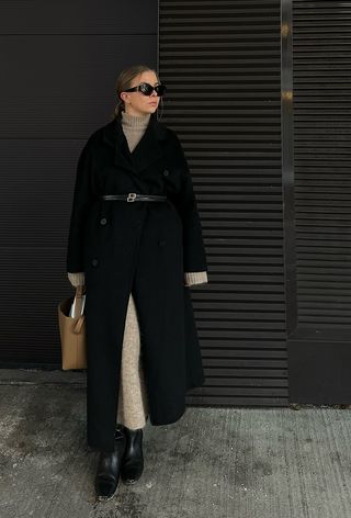 The winter capsule wardrobe item, the sweater dress, is shown in a photo of a woman standing outside wearing a long black coat over a tan turtleneck sweater dress with a black belt, tan tote bag, and black knee-high boots