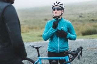 AlpKit Arro shell rain jacket being zipped up by a woman with a helmet on about to get on her bike