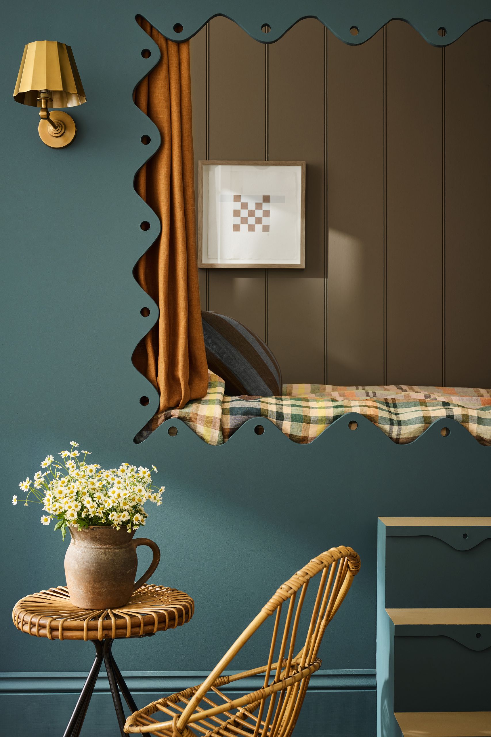 A cozy bed nook with brown paneling in the nook and plaid bedding. The wall outside the nook are teal and the window to nook is scalloped with an orange curtain. There is a rattan table and chair.