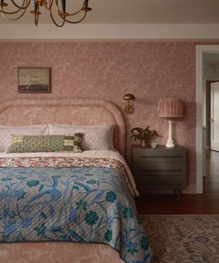 pink patterned drenched bedroom with matching wallpaper, headboard, lampshade, and bed skirt with a blue floral throw blanket at the foot of the bed