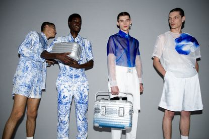 Four male models, two wearing white outfits with blue botanical patterns, and two wearing white and blue shirts with white trousers or shorts