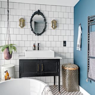 Bathroom with white bath and tiled wall, blue wall, grey and white tiled floor, black and white sink, hanging plant, wall lights and beige and black hamper