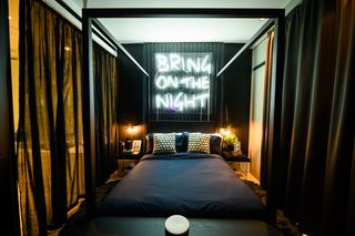 Black four poster bed with dark sheets, white cushions and bright white neon sign above headboard